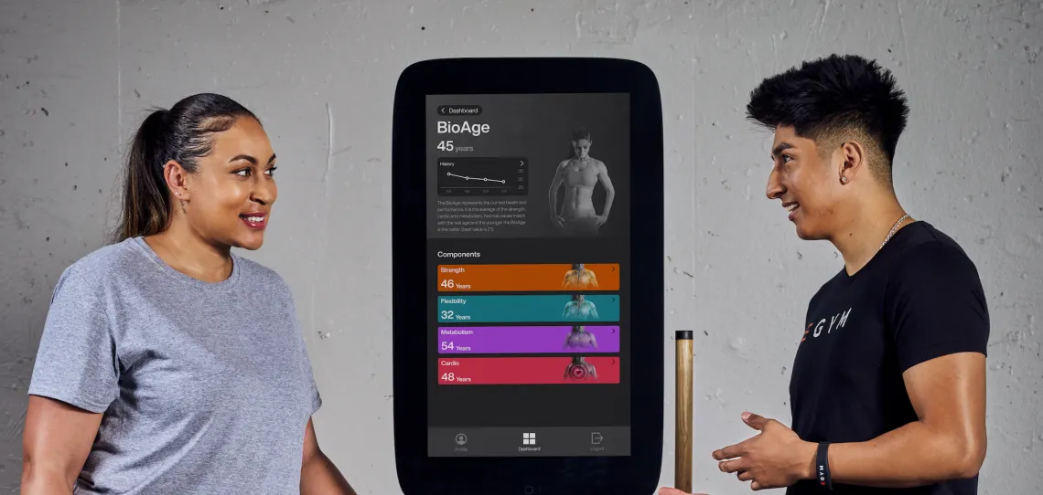 a woman looking at the egym display showing bioage 45 year old and an egym male trainer gesturing to the display that is between them.